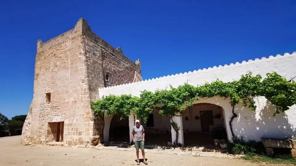 Torre de pedra quadrada vora una casa blanca amb parres