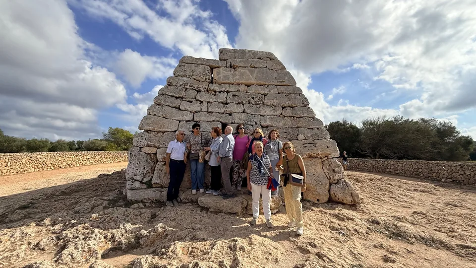 Besuchergruppe vor einer talayotischen Naveta aus Stein unter blauem Himmel mit Wolken