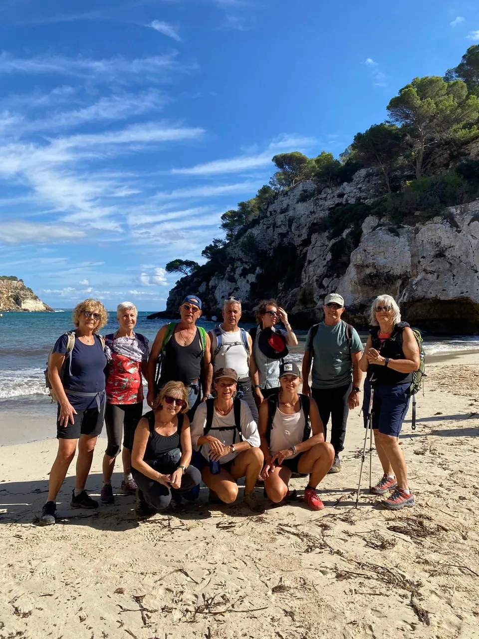 Grupo de diez excursionistas posando en la arena de una cala, con un acantilado y pinos al fondo